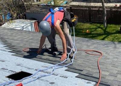 Professional roofer installing asphalt shingles on a residential roof in Regina, Saskatchewan, using a nail gun and wearing safety gear.