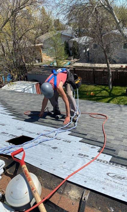 Professional roofer installing asphalt shingles on a residential roof in Regina, Saskatchewan, using a nail gun and wearing safety gear.